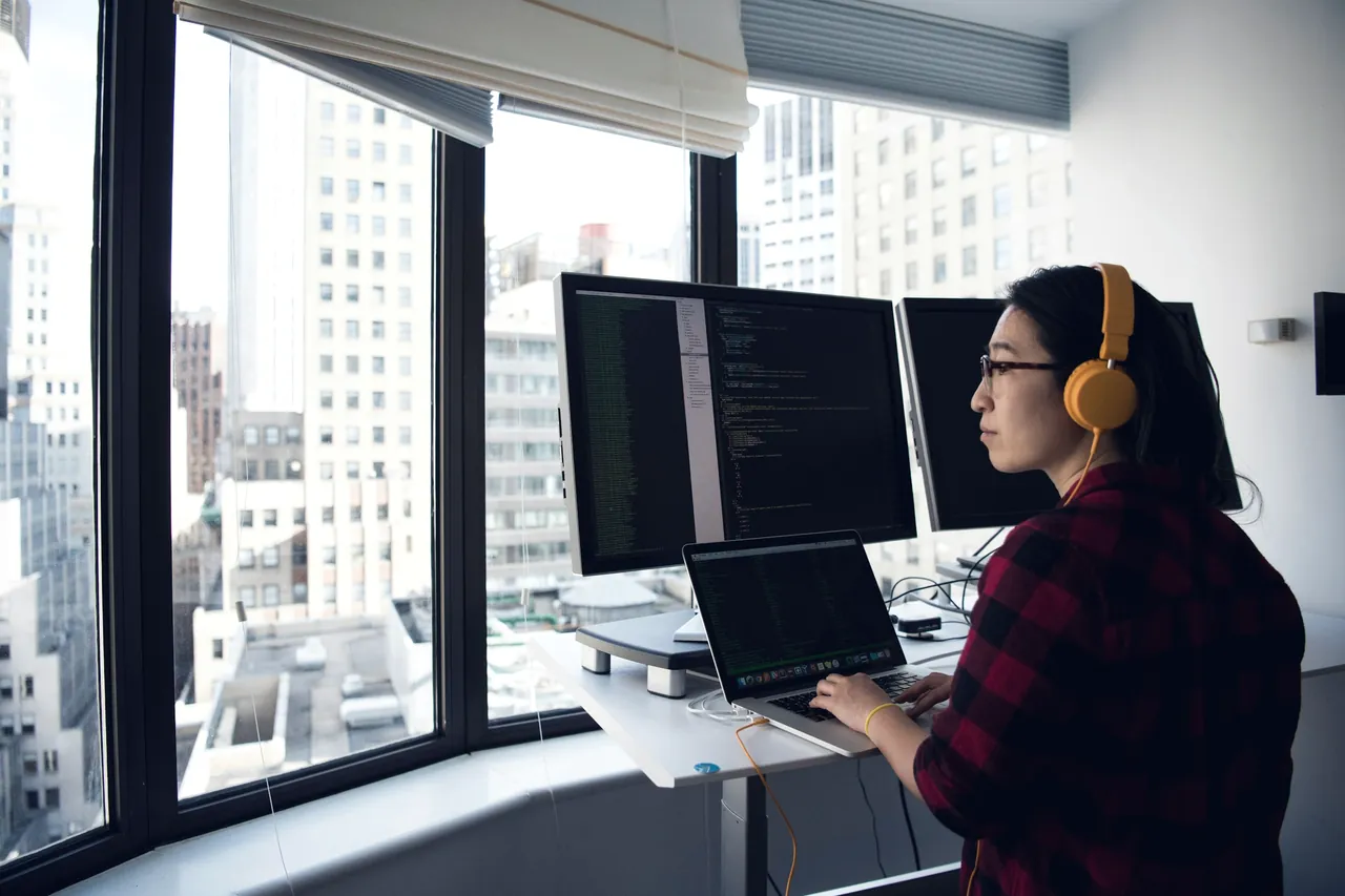 A woman wearing headphones works at two monitors beside a window